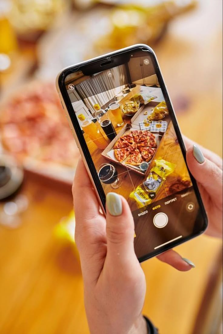 Person taking a photo of a pizza with a smartphone on a wooden table.