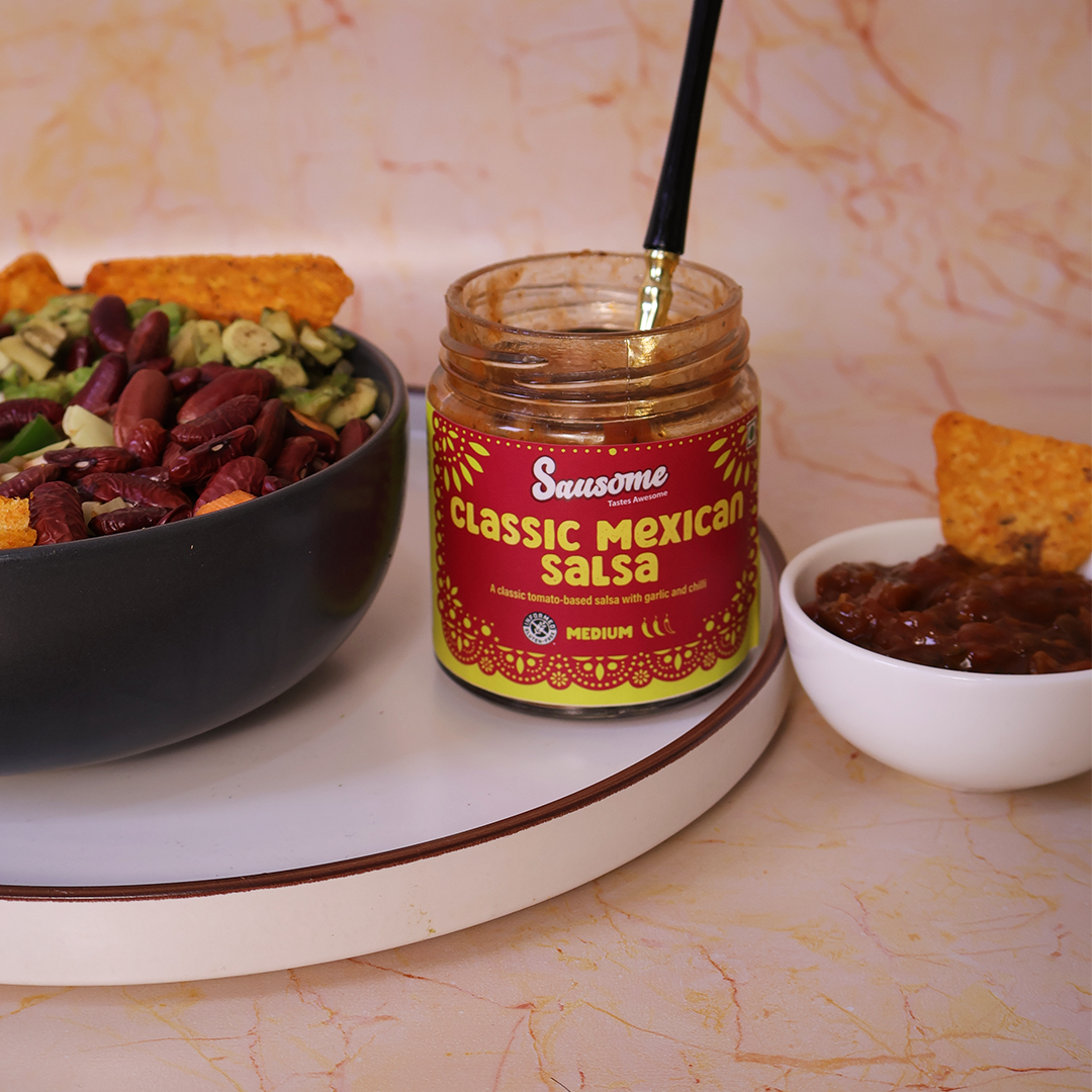 Jar of Sausome Classic Mexican Salsa with a bowl of guacamole and tortilla chips on a marble surface.