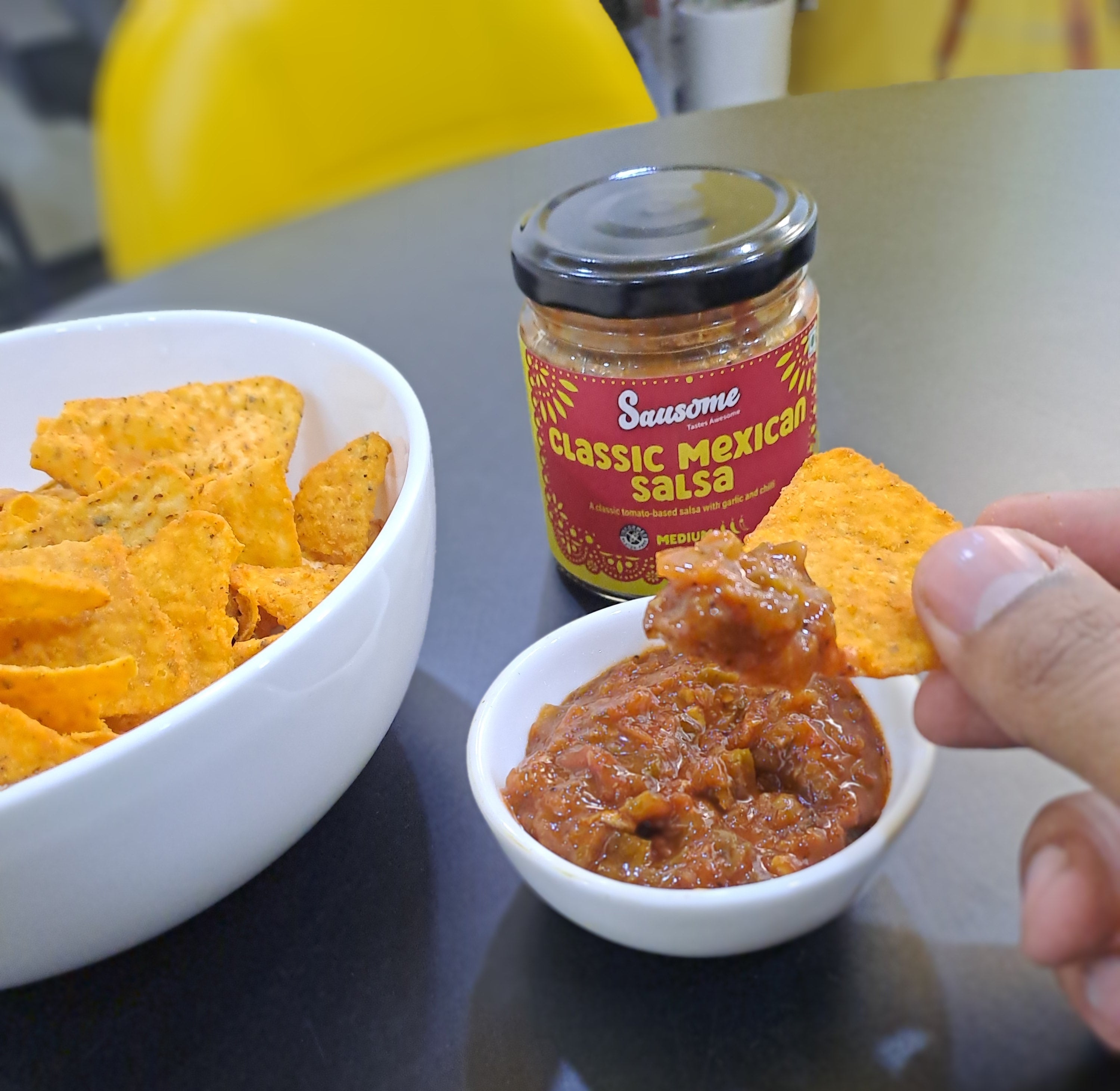 Person dipping a tortilla chip into a small bowl of salsa with a jar of salsa in the background.

