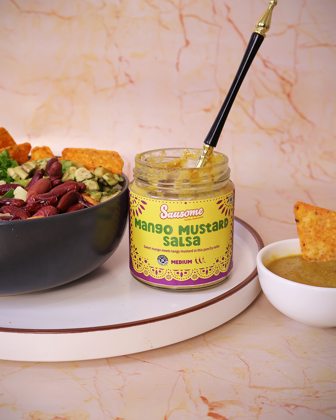 Jar of Sausome Mango Mustard Salsa with a bowl of guacamole and chips on a marble surface.