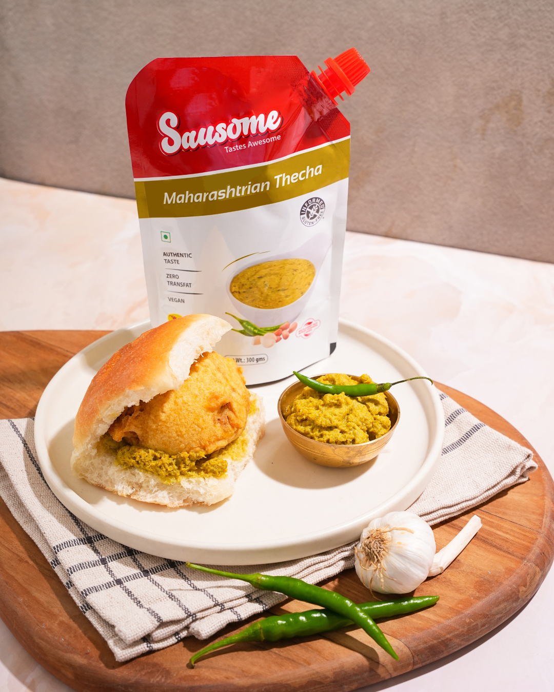 Sausome Maharashtrian Thecha packaging with a plate of Vada Pav on a wooden board.