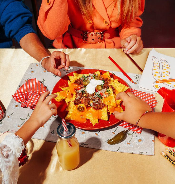 Two people reaching for a plate of nachos at a table with a bottle of orange juice.