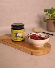 Jar of Thai chili oil and bowl of dried red chilies on a wooden board with a plant in the background.

